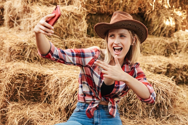 Vaqueira loira jovem e alegre sentada em um palheiro no celeiro, tirando uma selfie