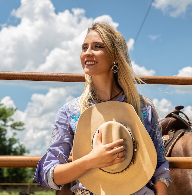 Vaquera rubia hermosa con el sombrero que se coloca cerca del fondo del rancho del caballo