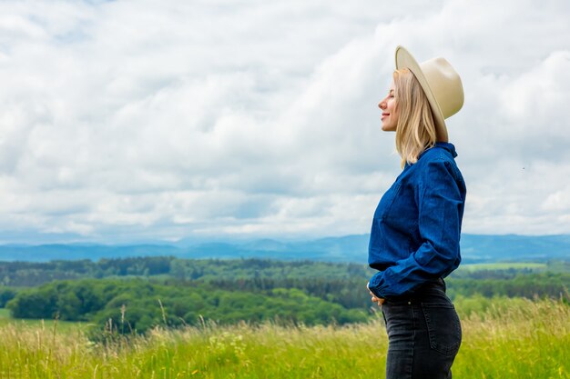 Vaquera rubia con sombrero en pradera con montañas