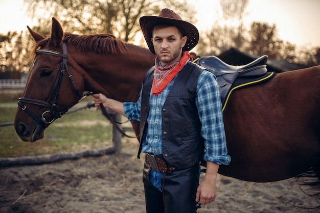 Foto vaquero brutal en jeans y chaqueta de cuero posa con caballo en el rancho de texas, occidental. persona del sexo masculino vintage con animal, salvaje oeste