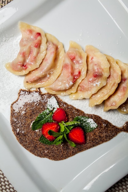 Vareniki con fresas, menta en un plato blanco grande, sobre un tapete de comida - fondo de estera de bambú Junto al cuchillo y tenedor - cubiertos. Cuaresma, menú vegetariano - vareniki con cerezas y salsa de fresa