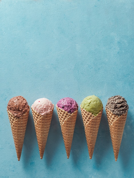 Varias bolas de helado en conos con espacio de copia. Colorido helado en conos de chocolate, fresa, arándano, pistacho o matcha, galletas galletas sandwich de chocolate sobre fondo azul. Vista superior