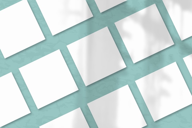 Várias folhas quadradas de papel texturizado branco contra uma parede azul esverdeada. A luz natural projeta sombras da janela. Camada plana, vista superior. Orientação horizontal