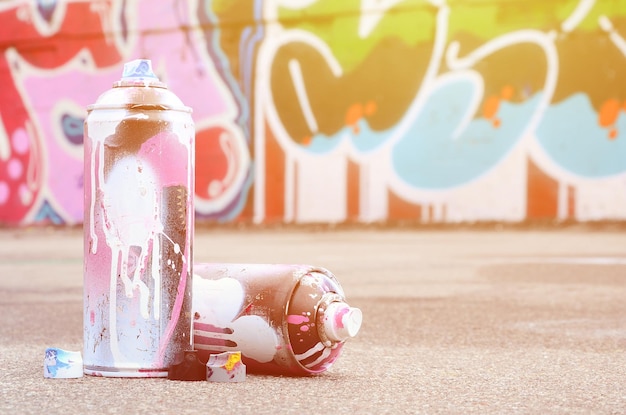 Várias latas de spray usadas com tinta rosa e branca e tampas para pulverizar tinta sob pressão estão no asfalto perto da parede pintada em desenhos coloridos de grafite