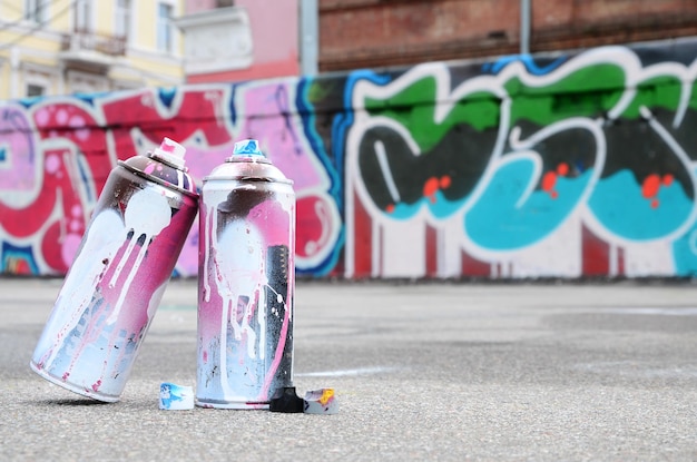 Várias latas de spray usadas com tinta rosa e branca e tampas para pulverizar tinta sob pressão estão no asfalto perto da parede pintada em desenhos coloridos de grafite