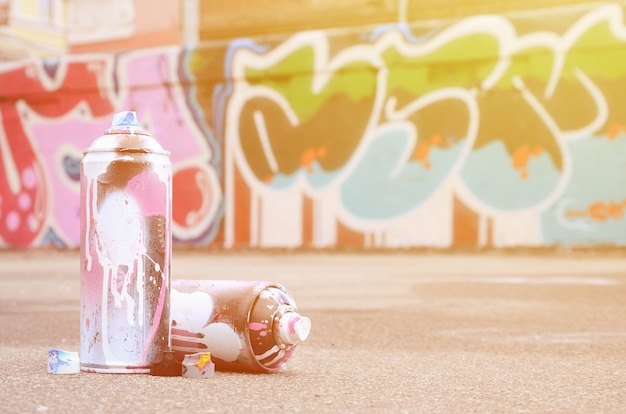 Várias latas de spray usadas com tinta rosa e branca perto da parede pintada em desenhos coloridos de grafite