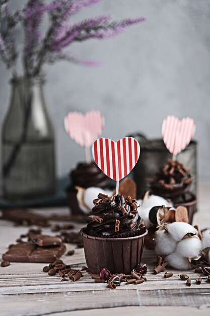 Varias magdalenas o cupcakes con crema en forma de chocolate en la mesa blanca. Una tarjeta en forma de corazón para el día de San Valentín. La mano de una mujer desmenuza el chocolate rallado en un pastel.