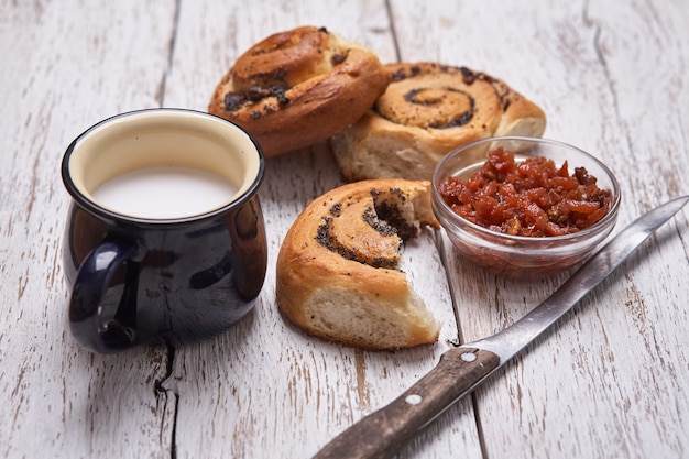 Variedad de bollos caseros de hojaldre de canela servidos con taza de leche, mermelada, mantequilla como desayuno sobre tabla blanca de mesa de madera