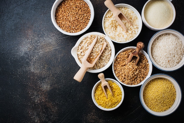 Variedad de cereales en copos de arroz, mijo, trigo sarraceno, avena. Superalimento en tazones de cerámica blanca sobre hormigón antiguo oscuro