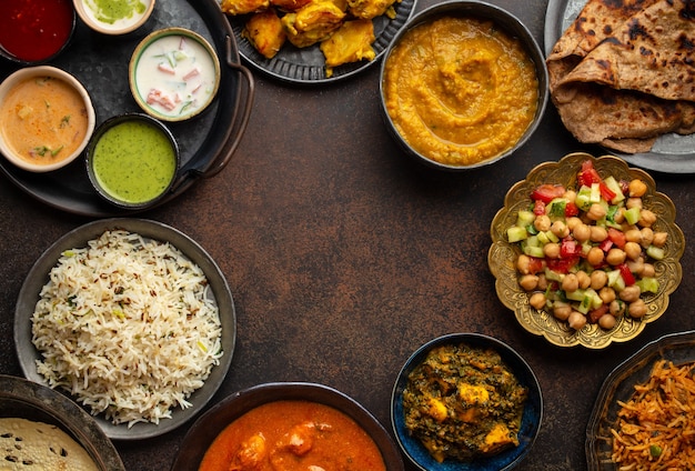 Variedad de comida india, diferentes platos y bocadillos sobre fondo rústico oscuro. Pilaf, pollo al curry con mantequilla, arroz, palak paneer, pollo tikka, sopa dal, pan naan, surtido de chutney. Copia espacio