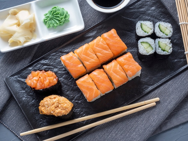 Una variedad de rollos y sushi gunkan anidados en un plato negro. al lado hay palitos de bambú y salsa de wasabi.