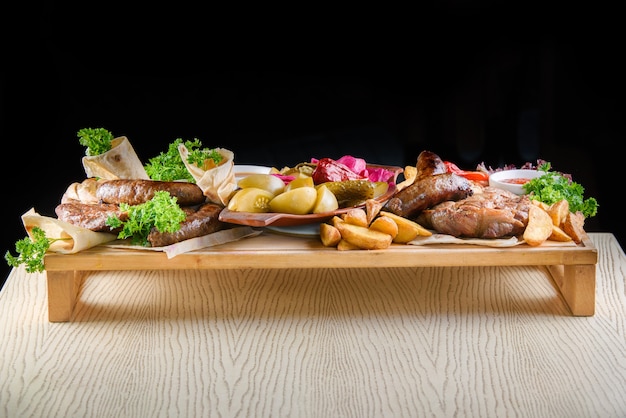 Variedade de carne frita, batata, salsicha, picles, tomate, pimentão, ervas, lavash em uma bandeja de madeira sobre a mesa.