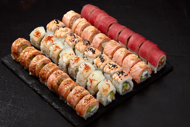 Variedade de diferentes tipos de rolos de sushi colocados na placa de pedra preta conjunto de sushi com pepinos salmão, cream cheese, abacate e sementes de gergelim imagem de sushi para menu
