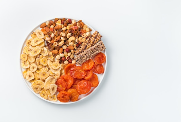 Variedade de frutas secas, nozes e barra de granola em um prato liso branco sobre uma parede clara. Vista superior com espaço de cópia. Lanches naturais e saudáveis.
