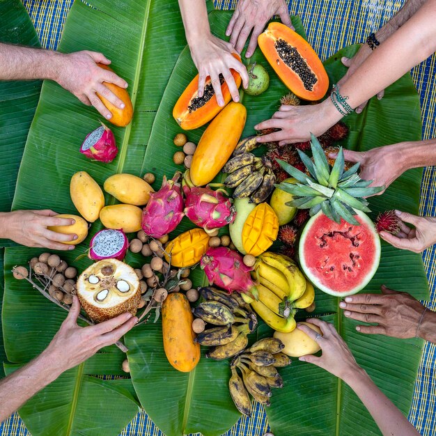 Variedade de frutas tropicais em folhas de banana verde e mãos de pessoas fecham sobremesa deliciosa fecham mango mamão pitaia e mãos vista superior