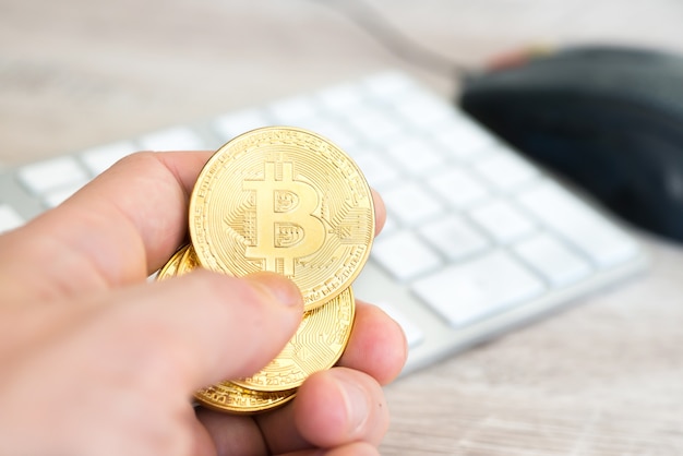 Varios bitcoins dorados en una mano cerca del teclado blanco y el mouse de la computadora. Concepto de minería de dinero electrónico