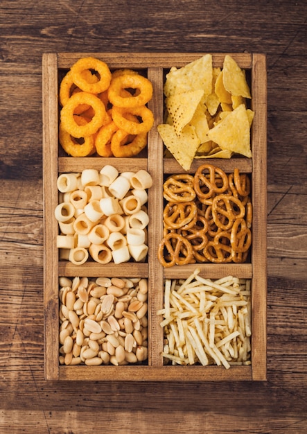 Varios bocadillos en caja de madera vintage sobre fondo de madera. Aros de cebolla, nachos, cacahuetes salados con palitos de patata y pretzels. Adecuado para cerveza y bebidas gaseosas.