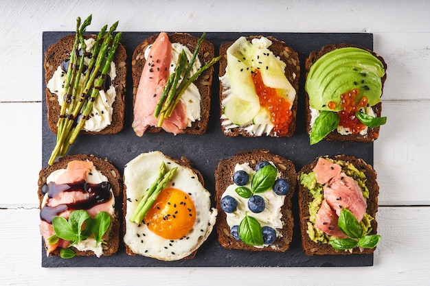 varios bocadillos saludables con diferentes rellenos. Pescado, caviar, aguacate, espárragos, pepino, hierbas, semillas de sésamo, pan sin gluten servido en una fuente oscura