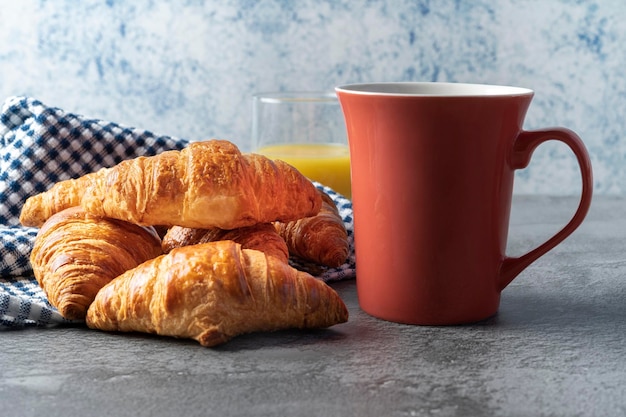 Vários croissants um em cima do outro em pano xadrez azul com uma xícara de café e suco de laranja