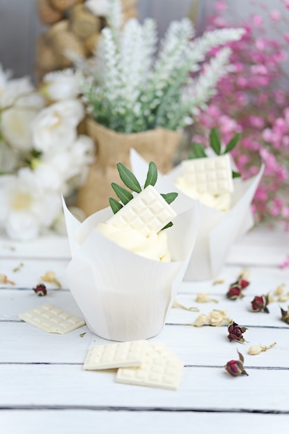 Varios cupcakes y muffins con crema de mantequilla blanca y una rosa vivaz sobre una mesa de madera blanca.