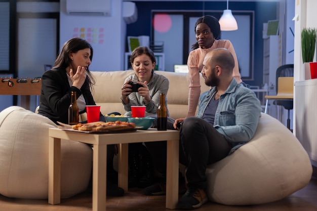 Vários grupos étnicos de colegas de trabalho jogam no console enquanto seguram o joystick. Amigos diversos alegres desfrutam de uma competição divertida no computador da tv enquanto tomam lanches e cerveja da mesa