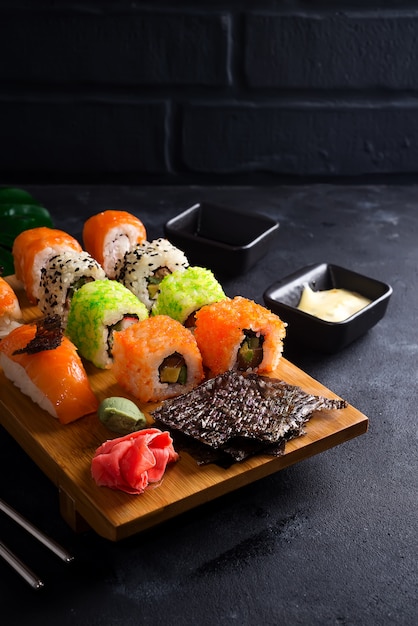 Varios juegos de sushi en placa de madera con palitos de pizarra, salsa y nori sobre negro