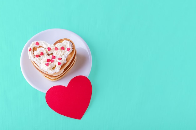 Varios panqueques caseros en forma de corazón cubiertos con crema con dulces coloridos en un plato blanco y una tarjeta de corazón rojo en blanco sobre un fondo azul brillante, copie el espacio. Concepto de desayuno de San Valentín