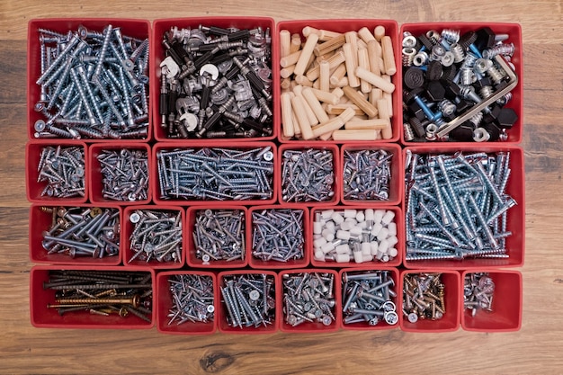 Varios tamaños y formas de tornillos, pernos, clavijas de madera, selección en una caja de bandeja de plástico roja sobre un fondo de madera, kit de montaje para muebles, vista superior