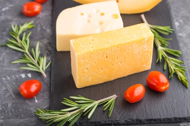 Vários tipos de queijo com alecrim e tomate na placa de ardósia preta sobre um fundo preto de concreto. Vista lateral, close-up, foco seletivo.