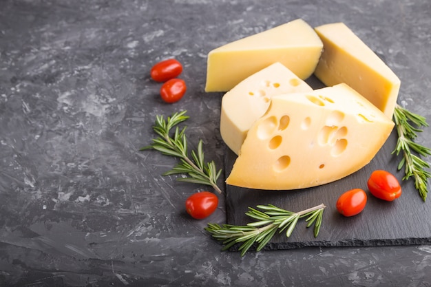 Varios tipos de queso con romero y tomates en pizarra negra sobre una superficie de hormigón negro. Vista lateral, copia espacio.