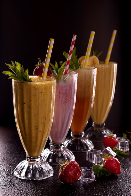 Varios vasos con papas fritas refrescantes con plátano, fresa y papaya, con hielo con tubo de cóctel sobre fondo negro