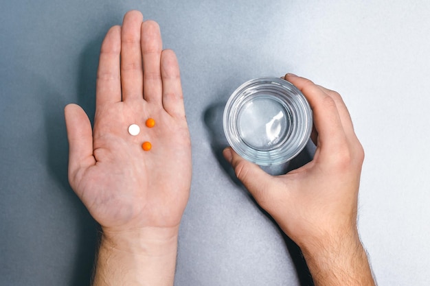 Varón caucásico con dos pastillas naranjas y una blanca en la palma izquierda y un vaso de agua en la mano derecha