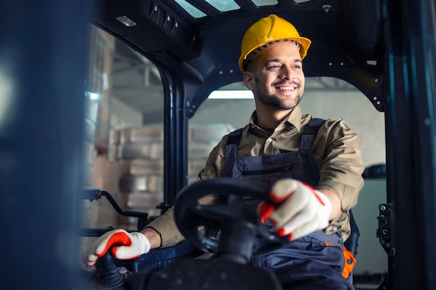 Varón caucásico joven en uniforme de trabajo y casco amarillo operando la máquina montacargas en la sala de almacenamiento del almacén.