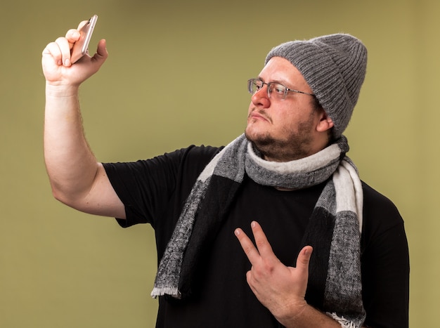 Varón enfermo de mediana edad con bufanda y gorro de invierno tomar un selfie mostrando gesto de paz