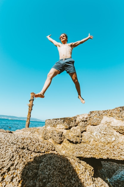 Varón joven con cabello largo saltando en el aire, sin camisa durante un día soleado, concepto de espacio y libertad, vacaciones, imagen fantástica, día de playa