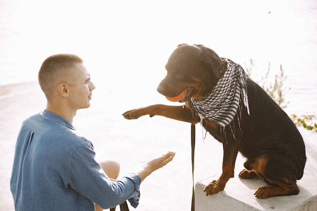 Varón joven guapo en traje casual jugando con lindo perro con bola naranja mientras está sentado cerca del lago. Perro da una pata