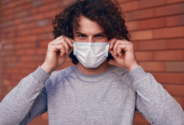 Foto varón joven con el pelo rizado mirando a la cámara y poniéndose una mascarilla médica mientras está de pie sobre el fondo borroso de la pared de ladrillo, durante la pandemia de coronavirus en la ciudad