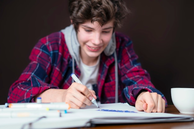 Varón joven sentado en la mesa y hacer dibujos en su cuaderno con marcador b