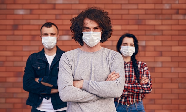 Varón joven serio con máscara médica cruzando los brazos y mirando a la cámara mientras está de pie cerca de amigos contra la pared de ladrillo durante la epidemia de coronavirus