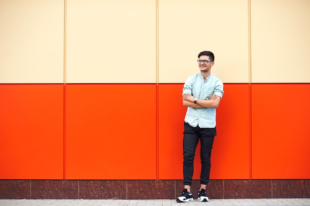 Varón joven sonriente con anteojos de pie contra la enorme pared naranja