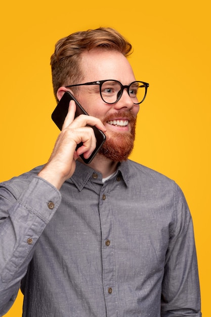 Varón joven sonriente en camisa gris y gafas conversando en el teléfono inteligente mirando a otro lado sobre fondo amarillo