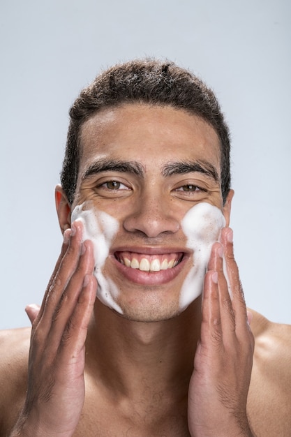 Varón joven sonriente extendiendo el lavado facial en sus mejillas