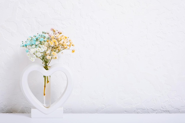 Vase Herzform mit mehrfarbigen Gypsophila-Blumen auf Weiß Minimalistische Inneneinrichtung mit Kopierraum