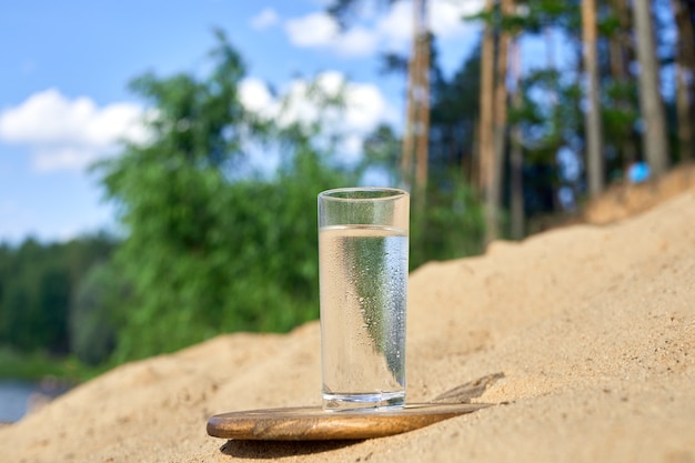 Vaso de agua fría sobre tabla de madera. Arena y árboles con fondo de naturaleza.