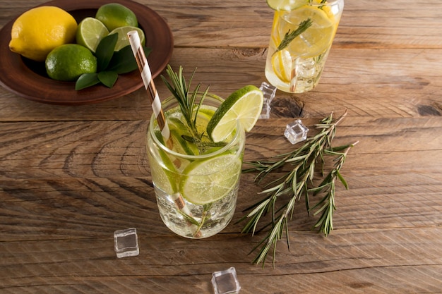 Un vaso de agua de lima, cubitos de hielo y romero, una refrescante bebida de verano, fondo marrón de madera con frutas y cubitos de hielo.