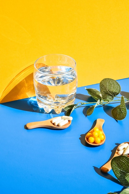 Vaso de agua y pastillas en una cuchara de madera sobre fondo azul y amarillo con luz dura y sombras