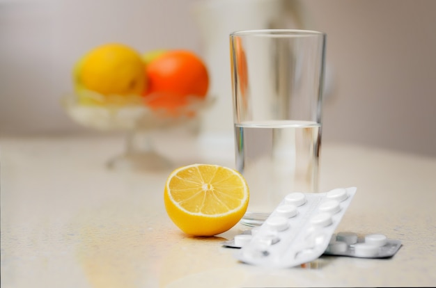 Un vaso de agua, pastillas para la enfermedad y un limón sobre la mesa con el telón de fondo de la fruta.