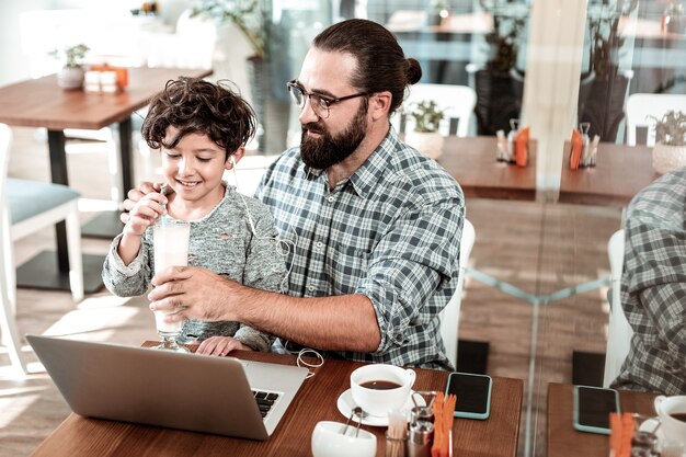 Foto vaso de batido. cuidado padre barbudo dando vaso de batido a su pequeño y lindo hijo pasar tiempo en la cafetería