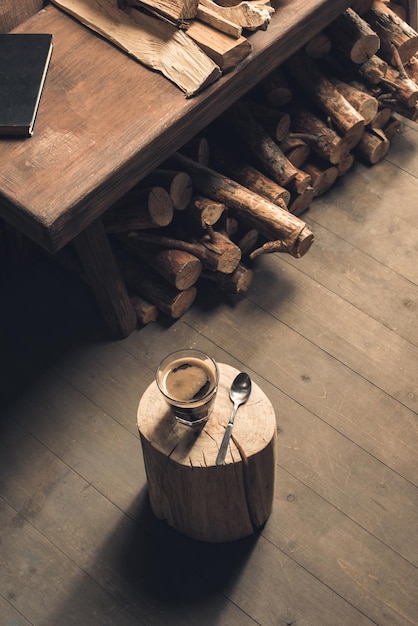 Vaso de bebida de café y cuchara en troncos de madera en el piso cerca de la mesa cerca de leña