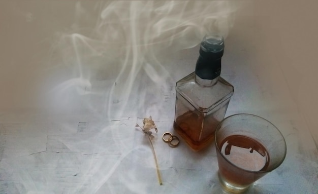 Vaso de botella de whisky y anillos de boda en humo sobre fondo gris